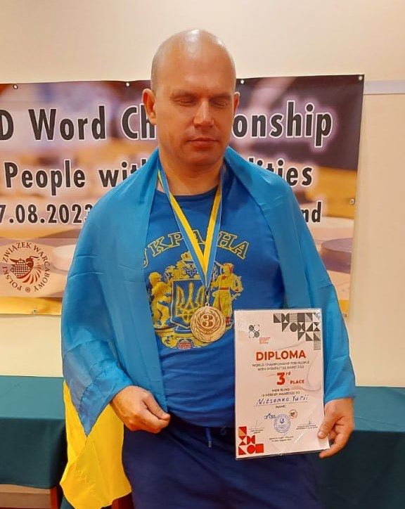 Лисий чоловік із прикритими очима, одягнений у синю футболку із золотим Тризубом, на плечі якого накинутий синьо-жовтий український прапор. На шиї спортсмена висить велика медаль, а в руках він тримає диплом, де чітко видно написи: DIPLOMA, 3rd Place (3-тє місце) та прізвище за участь у Всесвітньому чемпіонаті для людей з інвалідністю.