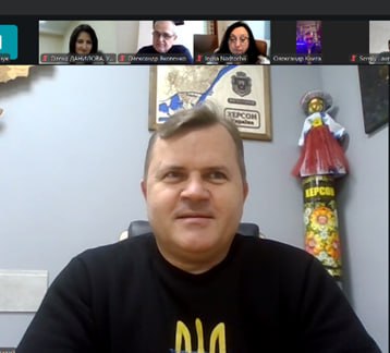 Чоловік у чорному світшоті з жовто-синім українським гербом (тризубом) на тлі відеоконференції Zoom з кількома учасниками. На стіні за ним висить карта Херсонської області.