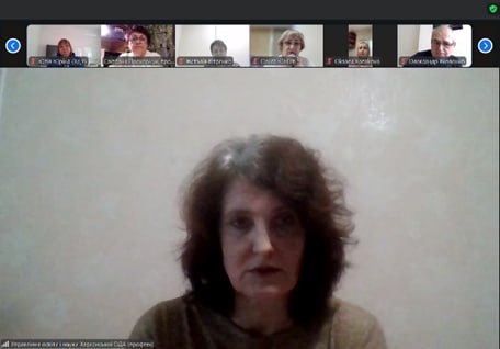 Жінка з темним хвилястим волоссям дивиться прямо в камеру, беручи участь у відеоконференції Zoom. Вона є головним доповідачем, а вище на екрані видно п'ятьох інших учасників.