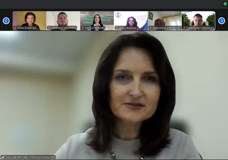 Жінка з темно-коричневим волоссям та намистом дивиться прямо в камеру, беручи участь у відеоконференції Zoom. Вона є головним доповідачем, а вище на екрані видно шістьох інших учасників.