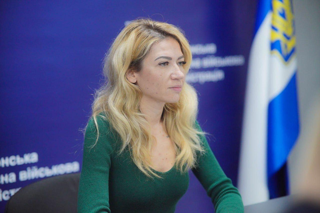 Жінка зі світлим, хвилястим волоссям у зеленому светрі сидить, дивлячись праворуч, на тлі синього банера з частково видимим текстом: Херсонська обласна військова адміністрація та елементами прапора.