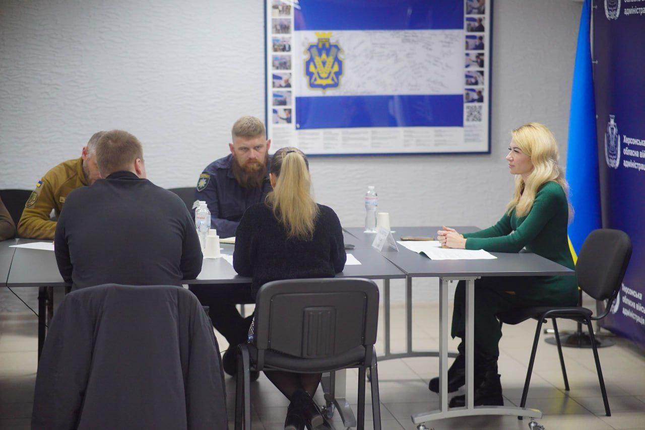 Четверо людей (троє чоловіків і одна жінка) сидять за столом під час зустрічі. На стіні позаду них висить великий плакат із прапором Херсонської області та підписами.