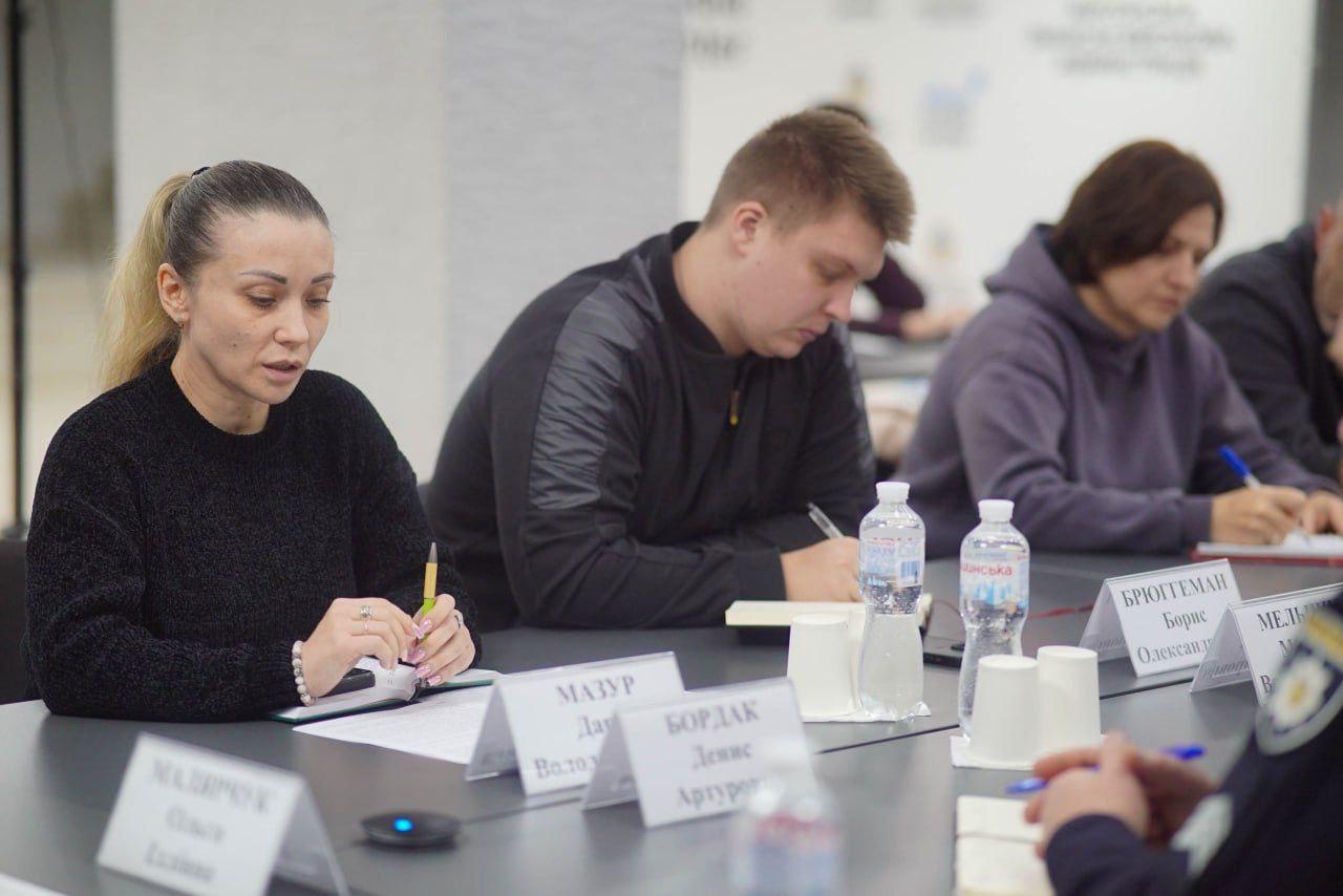 Троє людей (дві жінки та один чоловік) сидять за столом, записуючи нотатки під час зустрічі. На столі перед ними видно іменні таблички та пляшки з водою.