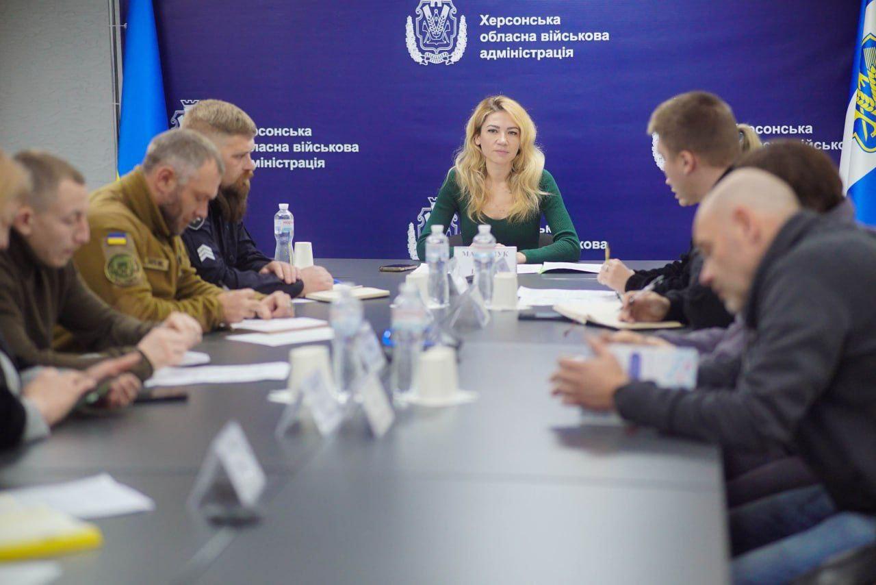 Жінка зі світлим волоссям у зеленому светрі сидить за столом на чолі зустрічі з кількома чоловіками та жінками в офіційному приміщенні з логотипом Херсонської обласної військової адміністрації на задньому плані.
