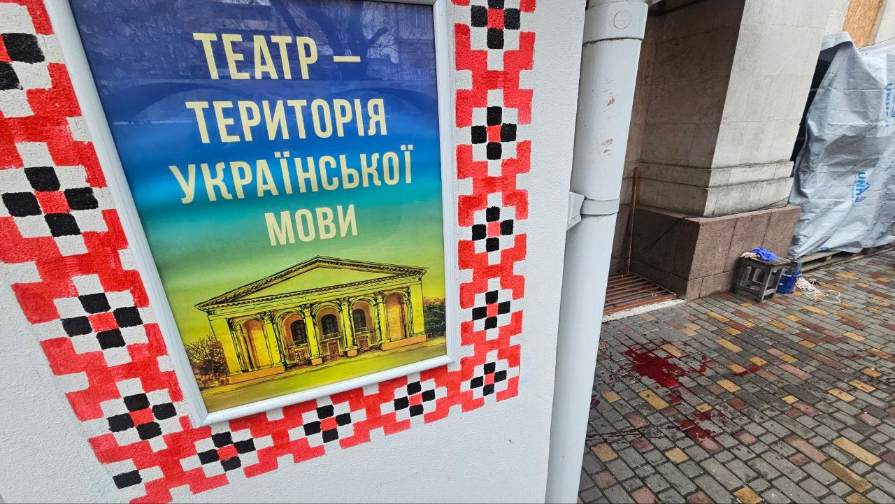 Плакат, оформлений в орнамент вишиванки, розміщений на стіні будівлі, містить напис: ТЕАТР – ТЕРИТОРІЯ УКРАЇНСЬКОЇ МОВИ та зображення фасаду театру. На бруківці поруч із плакатом видно плями, схожі на кров.
