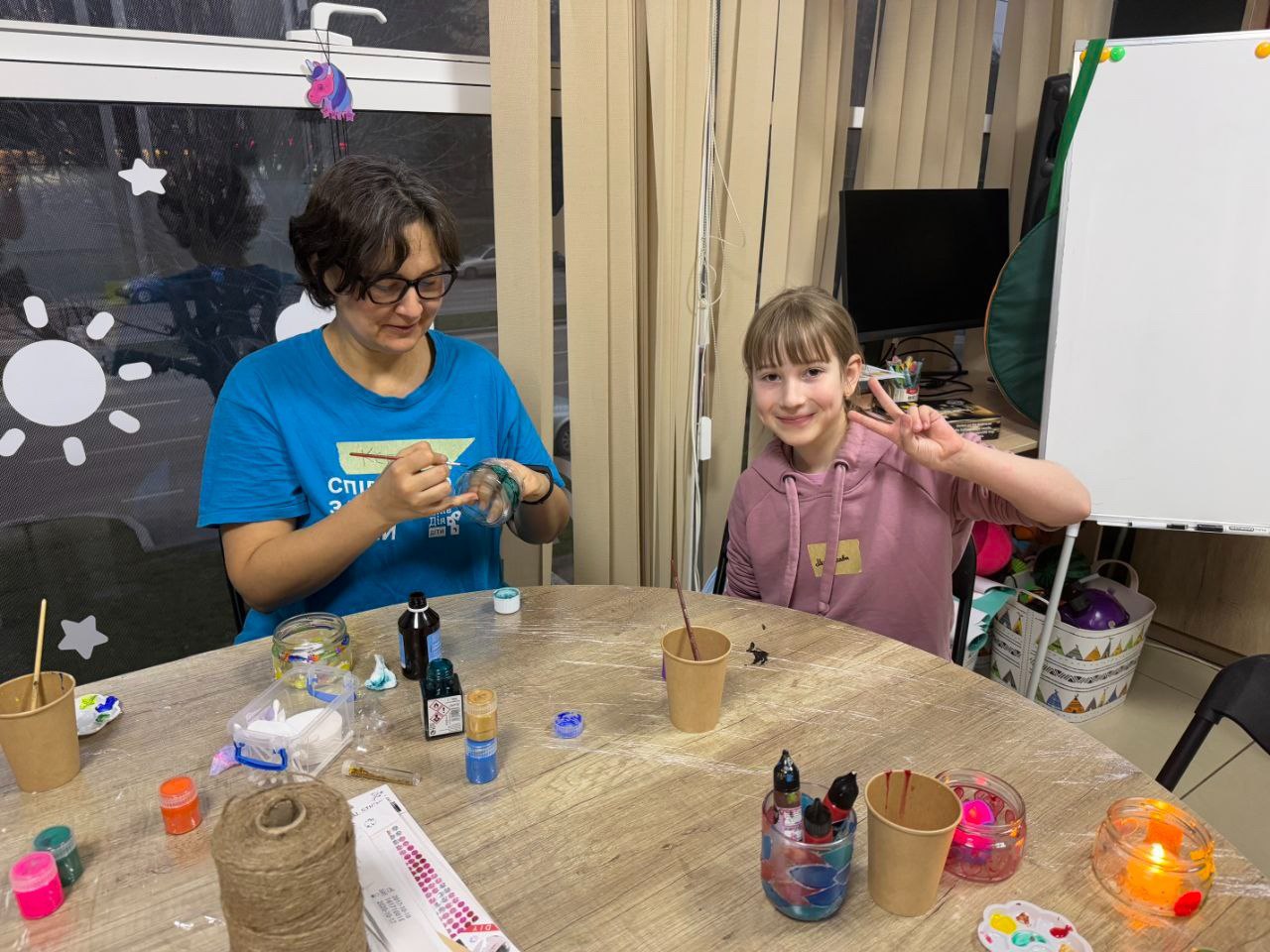 Жінка в синій футболці та дівчинка-підліток сидять за круглим столом, займаючись рукоділлям; дівчинка посміхається і показує знак V рукою.