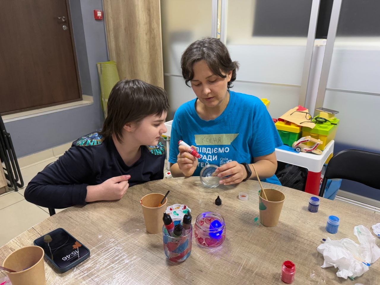 Жінка в синій футболці та дівчинка-підліток схилилися над круглим столом, займаючись рукоділлям. Жінка видавлює рідину з тюбика в маленьку прозору миску.