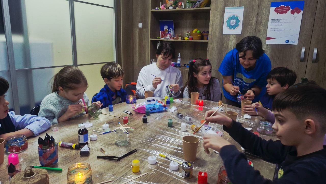 Круглий стіл, за яким сидять діти та дві жінки, зайняті спільним рукоділлям або творчим заняттям, прикрашаючи предмети.