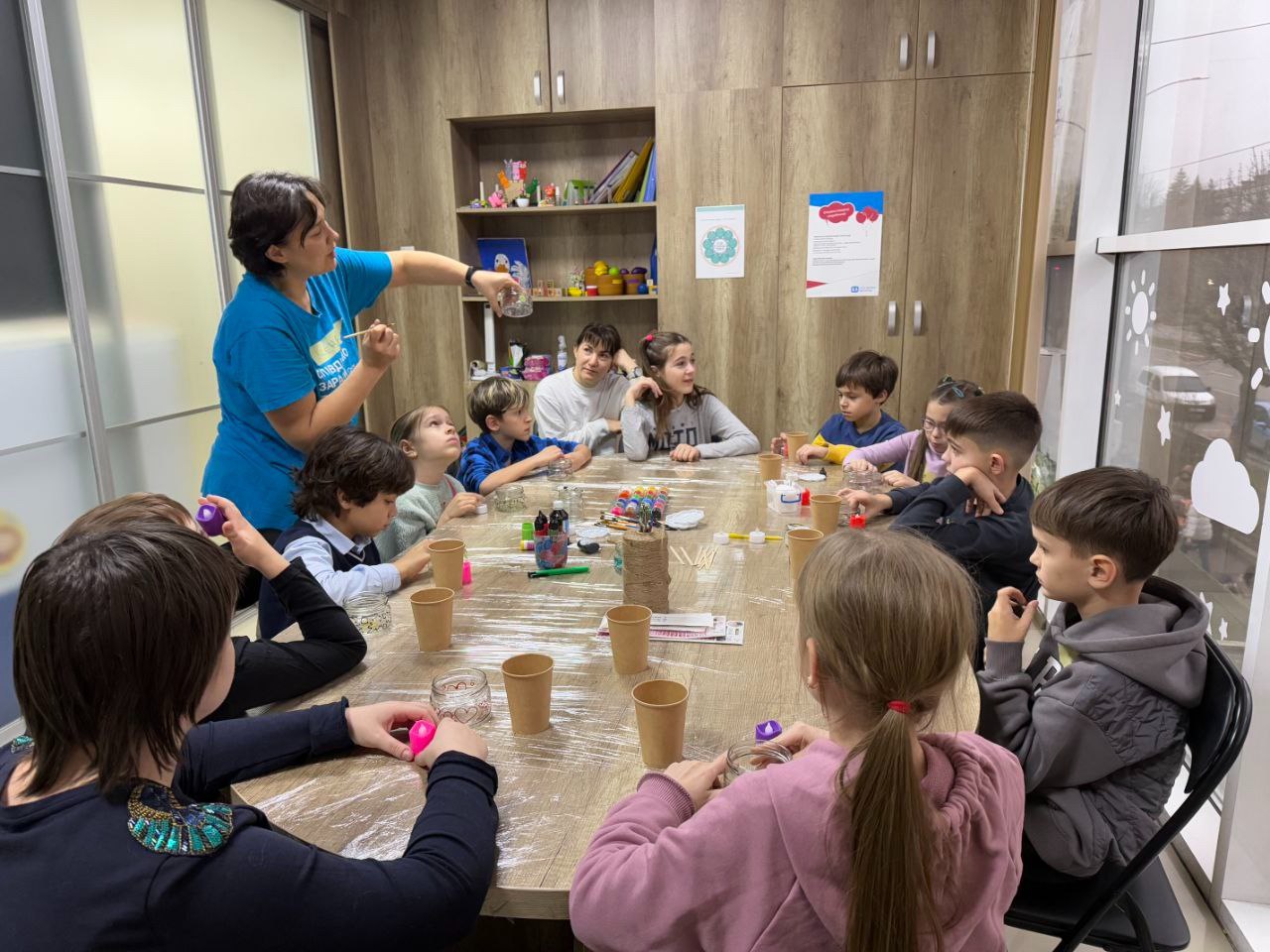 Група дітей сидить за столом, беручи участь у майстер-класі або занятті з рукоділля, яке проводить жінка в синій футболці, показуючи щось у скляній банці.