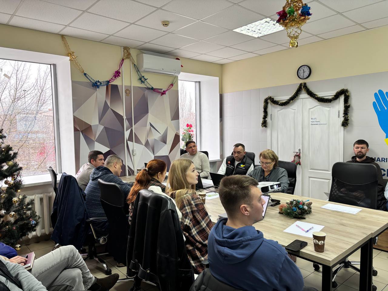 Група людей сидить навколо прямокутного столу, проводячи зустріч у кімнаті, прикрашеній різдвяною ялинкою та гірляндами, біля білих дверей.