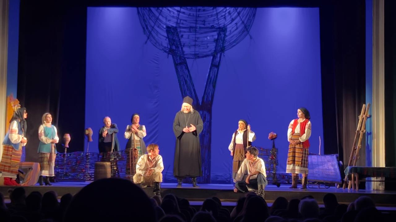 Сцена з театральної вистави. На сцені група акторів у традиційному українському та народному вбранні стоїть і сидить навколо чоловіка в чорній рясі (священника) на тлі яскраво-синього задника зі стилізованим плетеним деревом. Внизу видно силуети глядачів.