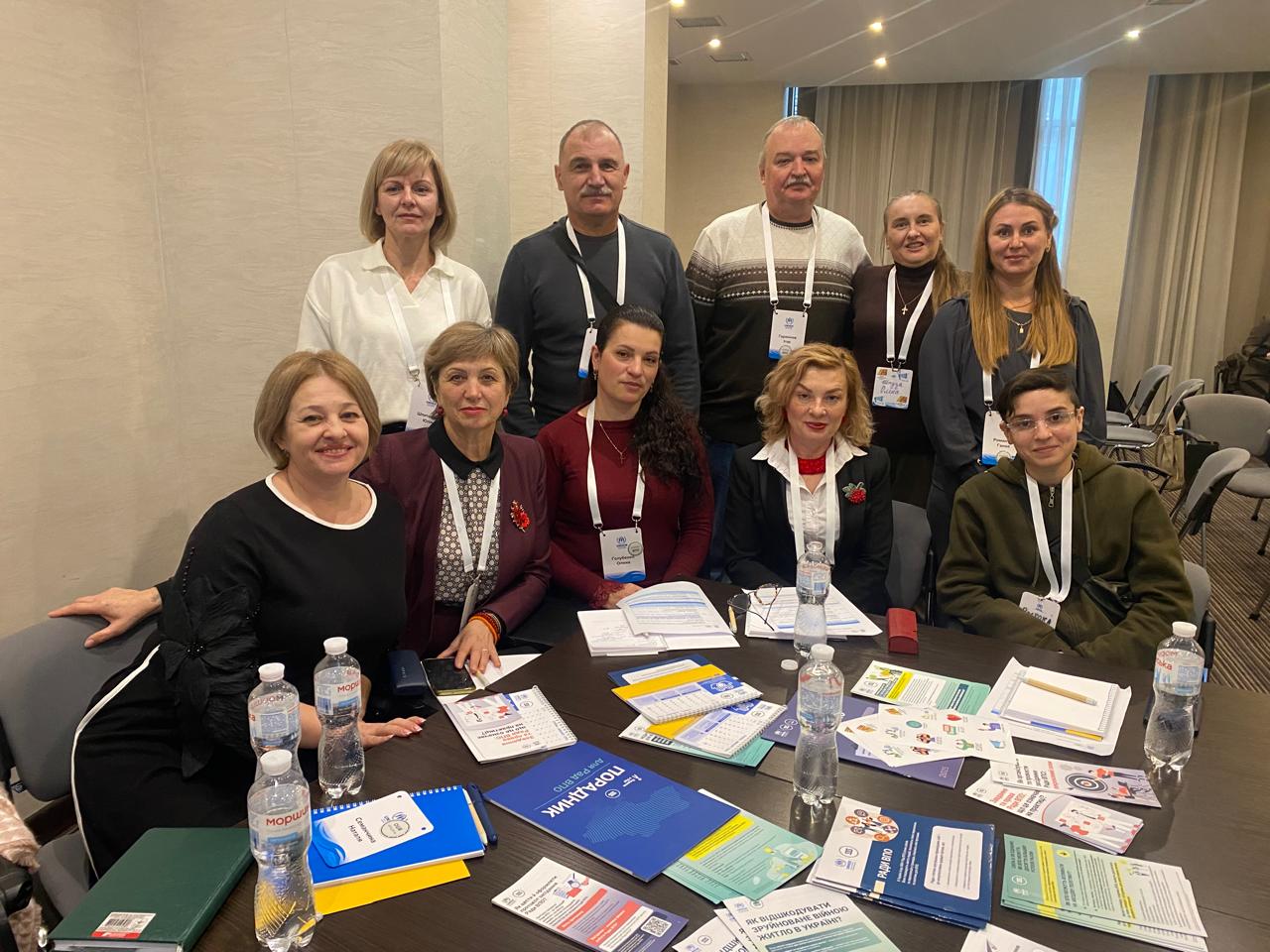 Група з дев'яти осіб (шість жінок і троє чоловіків) сидить за круглим столом, на якому лежать брошури та блокноти, і позує для фотографії на заході. Деякі люди мають ідентифікаційні бейджики на шиї.