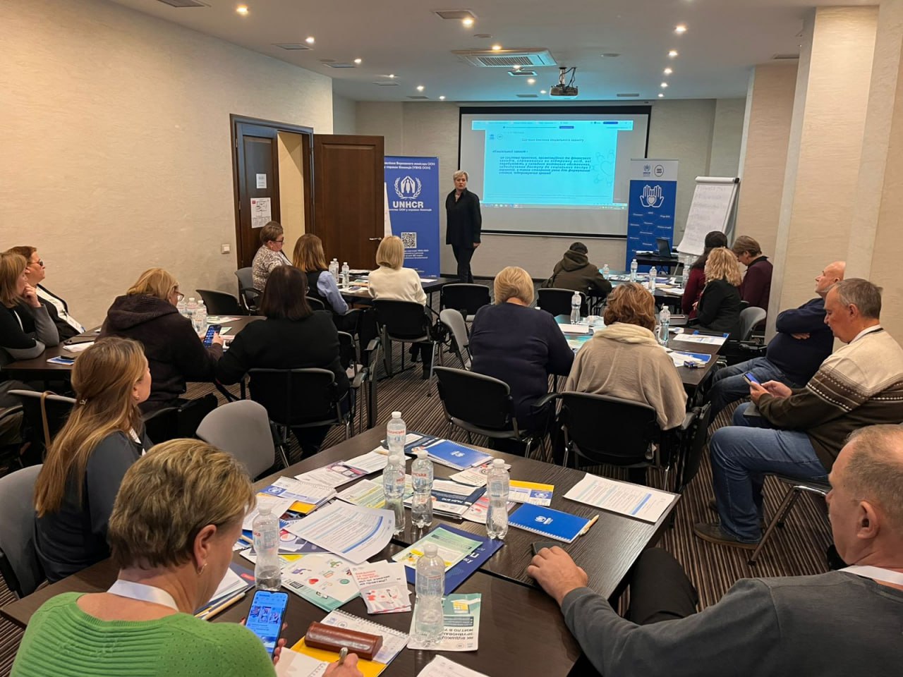 Загальний план семінару чи тренінгу в конференц-залі. Спікерка стоїть біля екрана з презентацією та логотипами UNHCR (УВКБ ООН) та інших організацій, звертаючись до аудиторії, що сидить за столами. На столах багато брошур, пляшок з водою та роздавальних матеріалів.