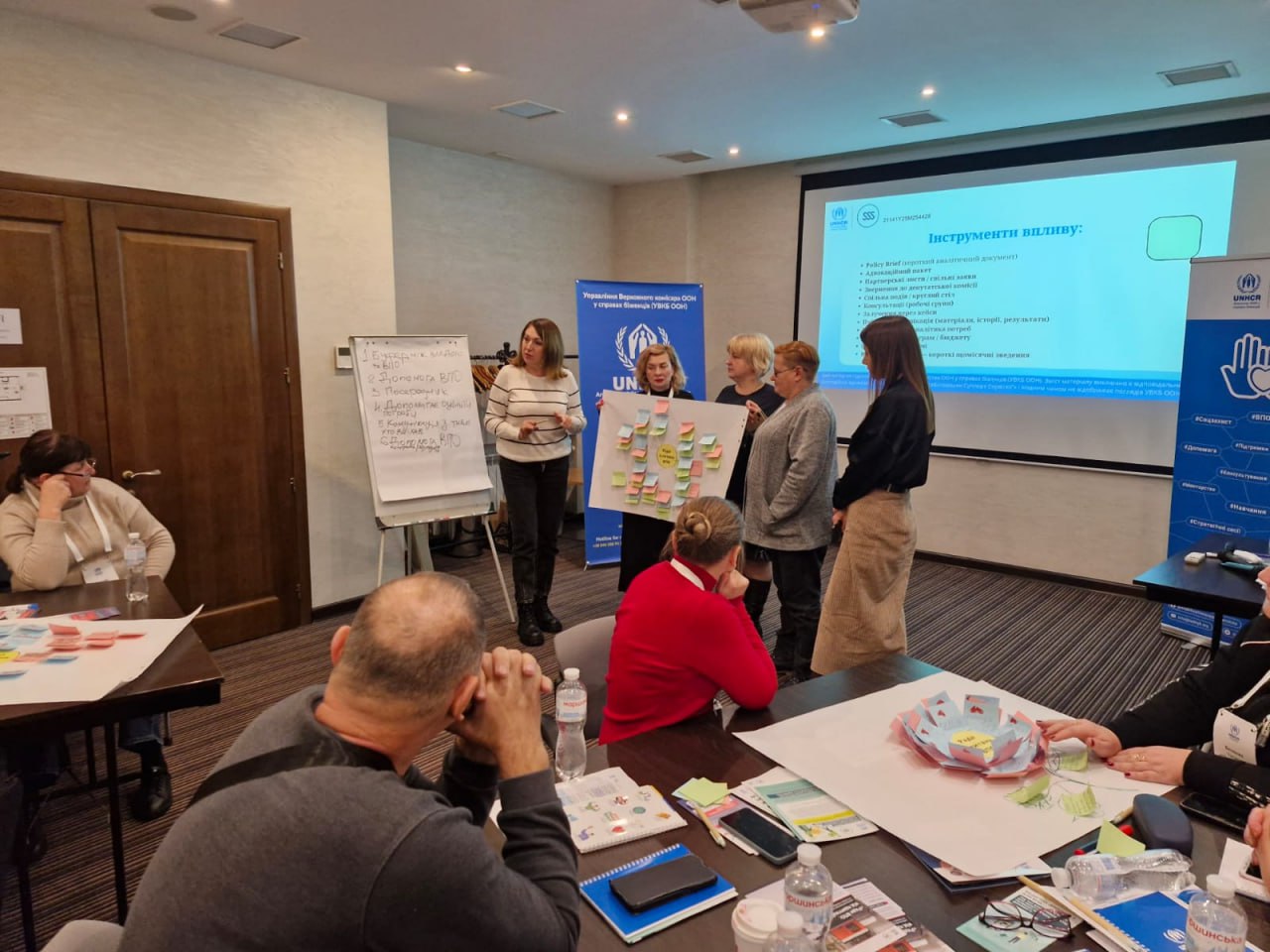 Чотири жінки стоять перед білою дошкою та тримають великий аркуш паперу з прикріпленими різнокольоровими стікерами-нотатками, демонструючи його групі людей, які сидять за столами під час семінару. На екрані проєктується слайд Інструменти впливу з логотипом UNHCR (УВКБ ООН).