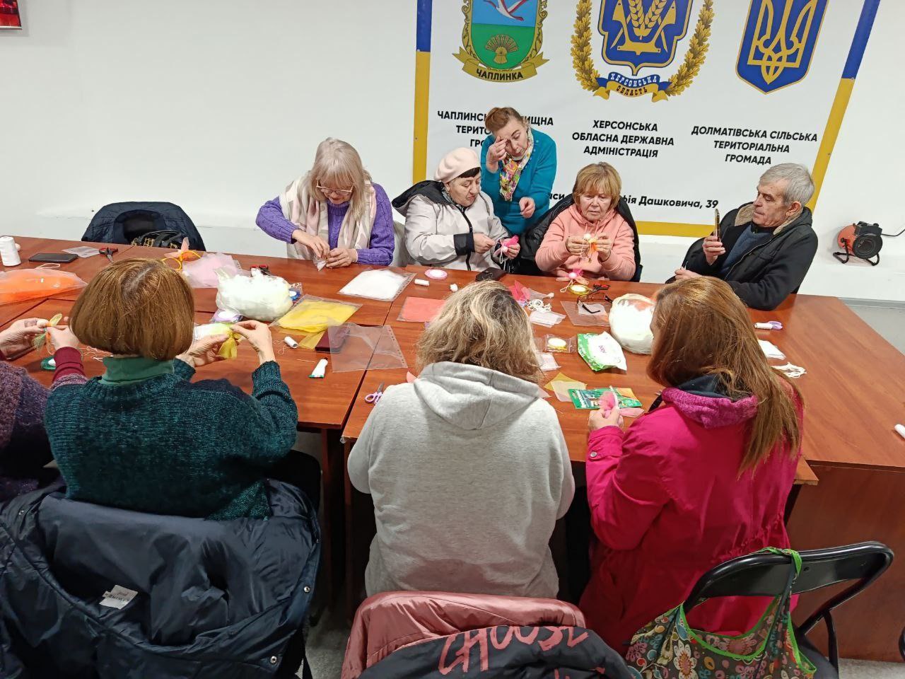 Групова фотографія, на якій приблизно дев'ять людей (переважно жінки старшого віку та один чоловік, який знімає на телефон) сидять за довгим коричневим столом і беруть участь у майстер-класі з рукоділля. На столі розкладені різні матеріали для творчості, включаючи тканини, нитки та клей. Жінки зосереджено працюють над своїми виробами.