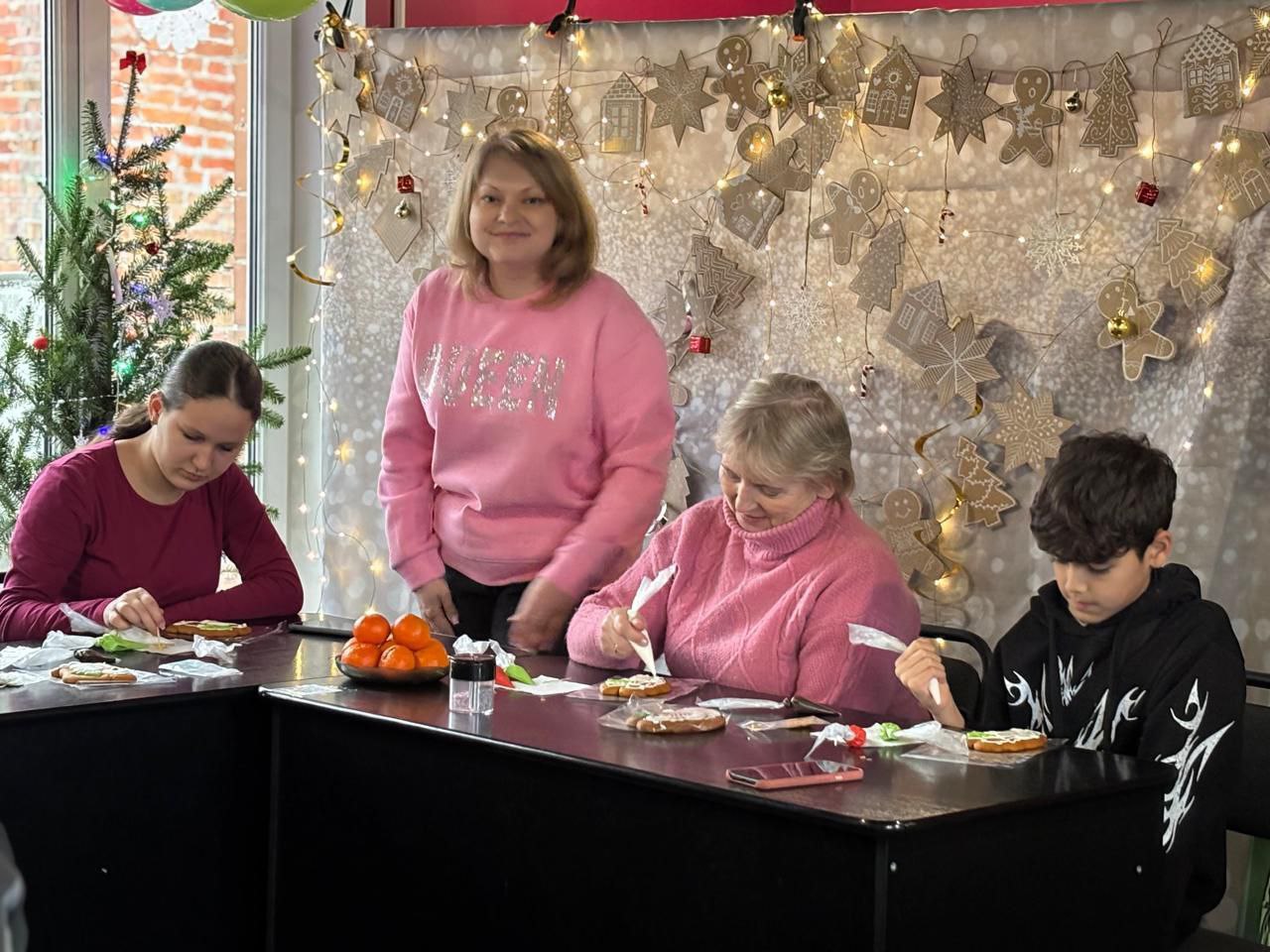 Чотири людини, серед яких двоє дорослих жінок у рожевих светрах та двоє підлітків, сидять за столом та прикрашають імбирні пряники. На тлі – різдвяна/новорічна декорація з гірлянд, ялинки та пряникових зірок.