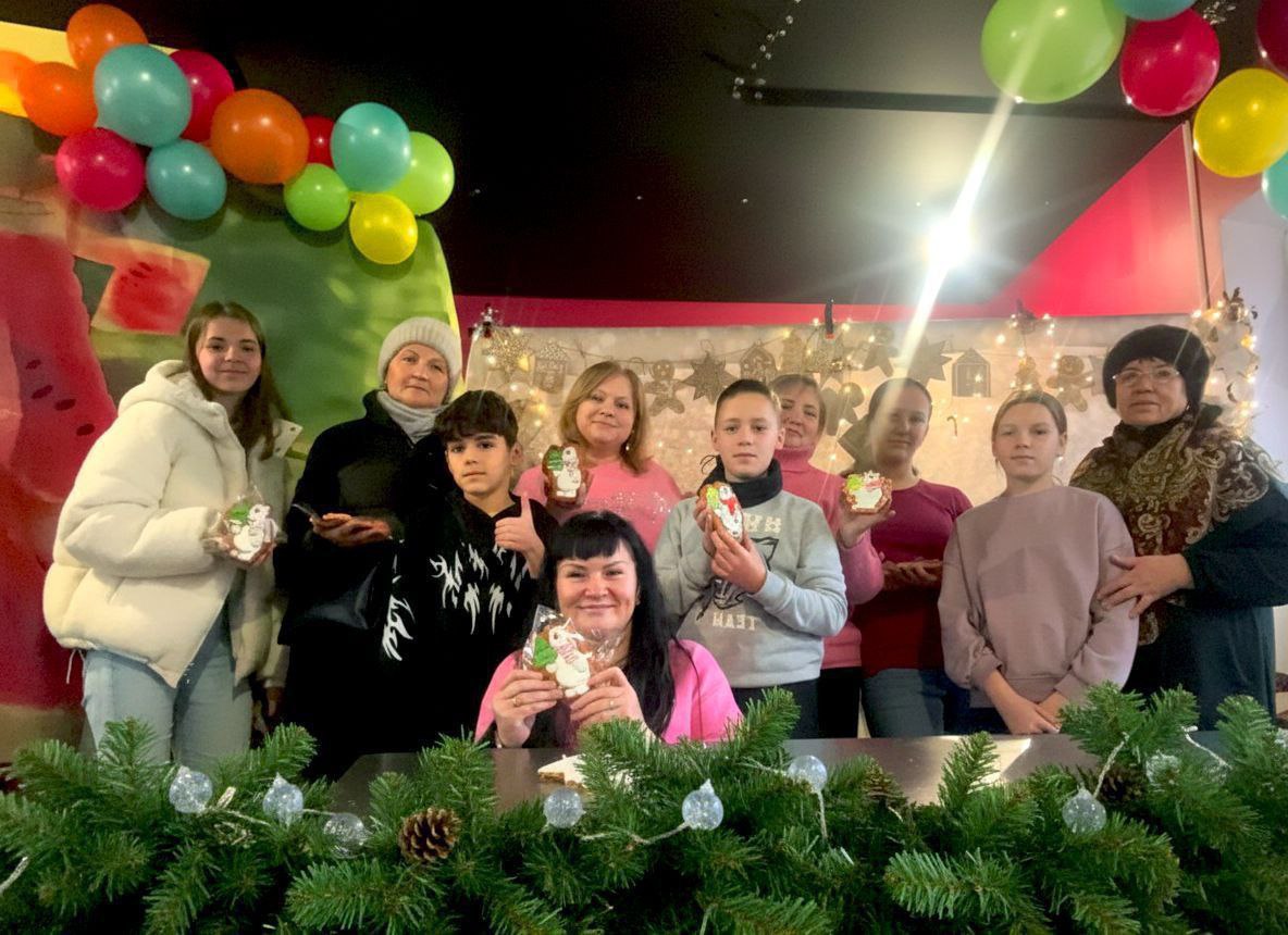 Група з деcяти людей – дорослих та підлітків – стоїть разом та посміхається, тримаючи в руках прикрашені імбирні пряники у формі сніговиків. На задньому плані видно святкові гірлянди, декорації та повітряні кульки, а на передньому плані – ялинову гірлянду.