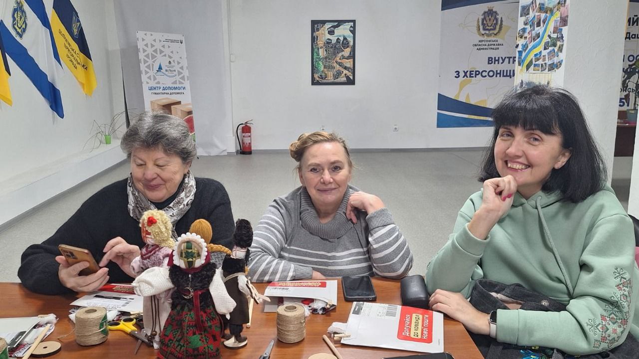 Три усміхнені жінки сидять за столом, на якому лежать нитки, матеріали для рукоділля та готові ляльки-мотанки. Одна жінка, яка ліворуч, дивиться у телефон.