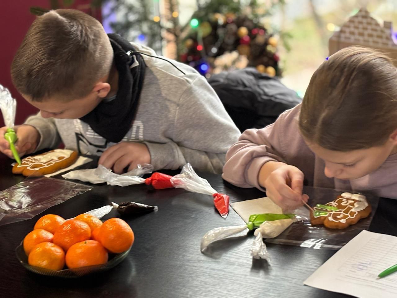 Двоє дітей (хлопчик і дівчинка) сидять за темним столом і зосереджено прикрашають імбирні пряники білою та зеленою глазур'ю. На столі лежать мандарини та кондитерські мішечки з глазур'ю. На задньому плані — святкова декорація в розфокусі.
