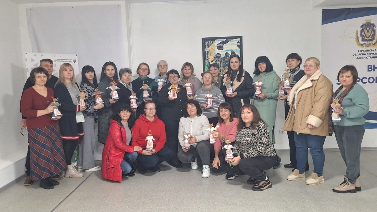 Групова фотографія приблизно двадцяти жінок, що стоять і присідають усередині світлого приміщення з білими стінами. Більшість із них тримають у руках яскраво прикрашені ляльки-мотанки. На задньому плані ліворуч видно чоловіка, який стоїть за жінками, а праворуч видно частину логотипу та напис українською мовою: ХЕРСОНСЬКА ОБЛАСНА ДЕРЖАВНА АДМІНІСТРАЦІЯ.