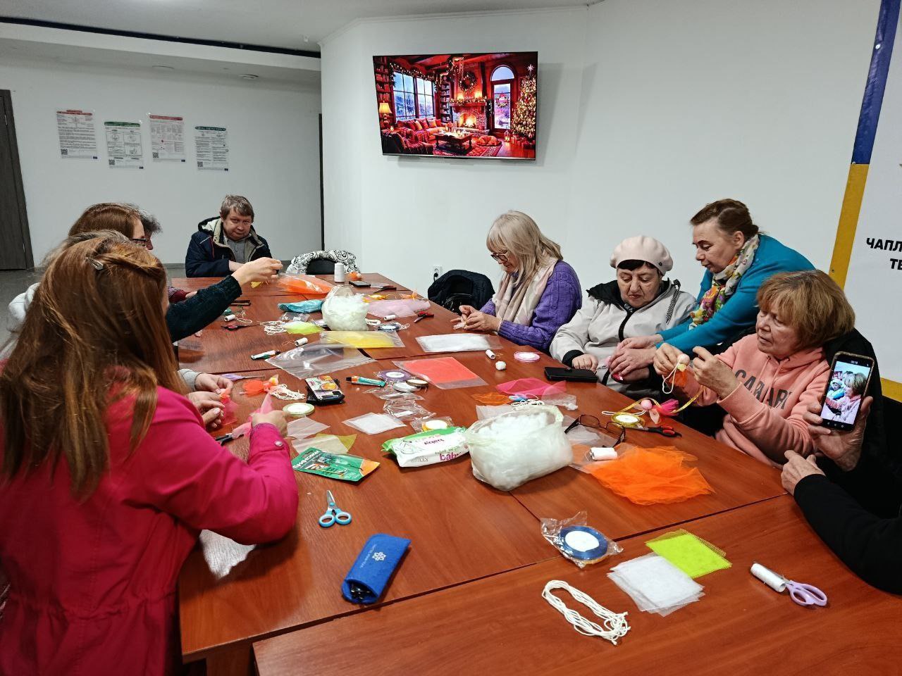 Група з дев'яти дорослих жінок та одного чоловіка сидять навколо великого дерев'яного столу, займаючись рукоділлям. Стіл заповнений різноманітними матеріалами для творчості, такими як кольорові тканини, стрічки, ножиці та наповнювач. Жінки одягнені у повсякденний одяг і зосереджено працюють руками, роблячи щось схоже на м'які іграшки чи прикраси. На стіні за ними висить великий телевізійний екран з зображенням затишної кімнати, прикрашеної до Різдва.