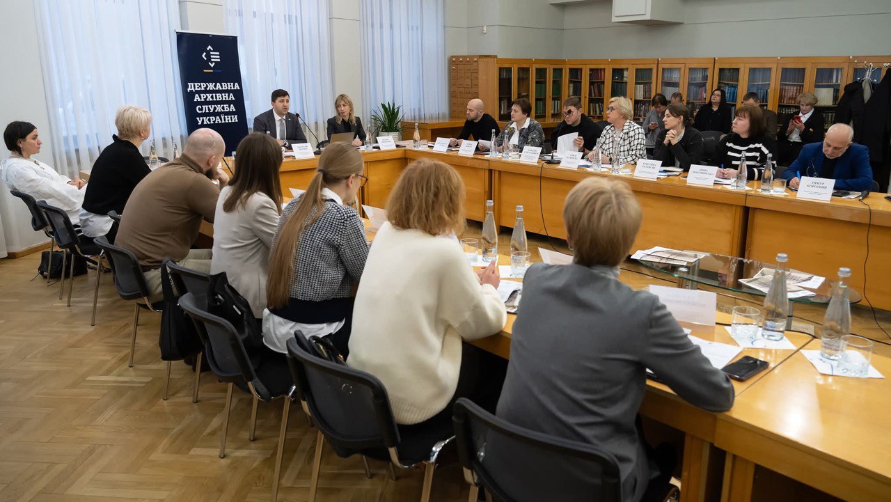 Група людей бере участь у нараді або конференції, що проходить у приміщенні з дерев'яними столами, де розміщено багато пляшок з водою та ідентифікаційні картки. Учасники сидять за довгим U-подібним столом. На задньому плані, на стіні, розміщений вертикальний синій банер з біло-жовтим логотипом Державної архівної служби України.