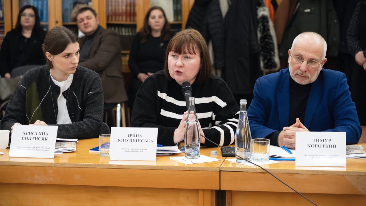 Троє людей — дві жінки та чоловік — сидять за довгим дерев'яним столом під час конференції або наради. Жінка в центрі, з мікрофоном і в чорно-білому смугастому светрі, говорить. На столі перед ними стоять пляшки з водою та склянки. На задньому плані видно книжкові полиці та присутніх слухачів.