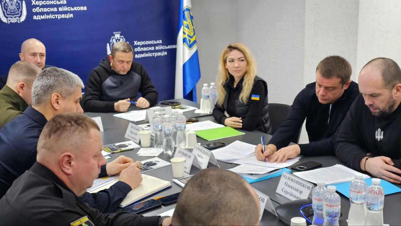 Велика група людей, що складається з чоловіків та жінок, сидить за довгим столом у конференц-залі. На столі багато пляшок з водою та документи. На задньому плані розташовані прапори України та Херсонської ОВА. Один з учасників активно щось записує.