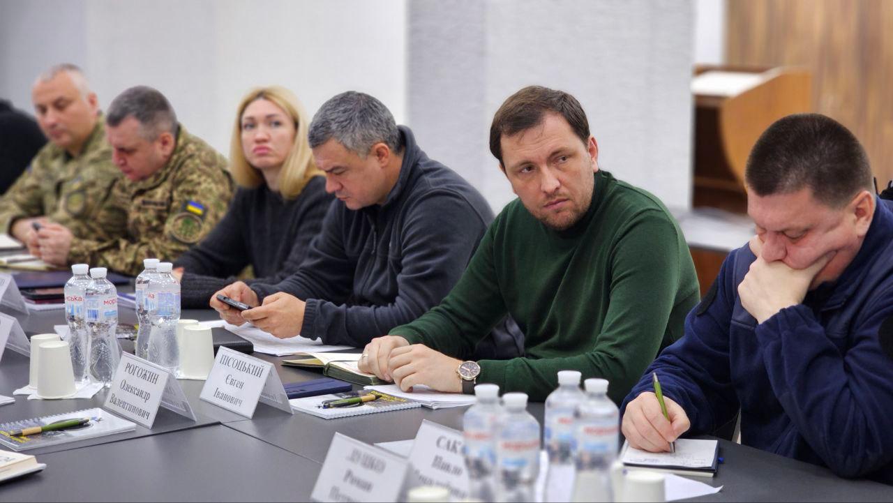 Група людей, (кілька чоловіків та жінка), сидять за довгим столом на зустрічі чи нараді. На столі видно пляшки з водою, блокноти, ручки та іменні картки. Чоловік у зеленому светрі в центрі дивиться прямо, а інший чоловік праворуч робить записи.