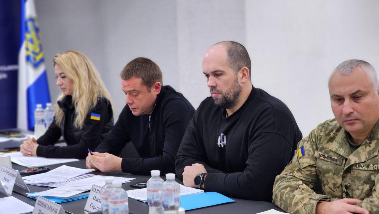 Четверо людей сидять за столом під час зустрічі. Троє в цивільному чорному одязі та один у військовій формі Збройних Сил України (зліва направо).