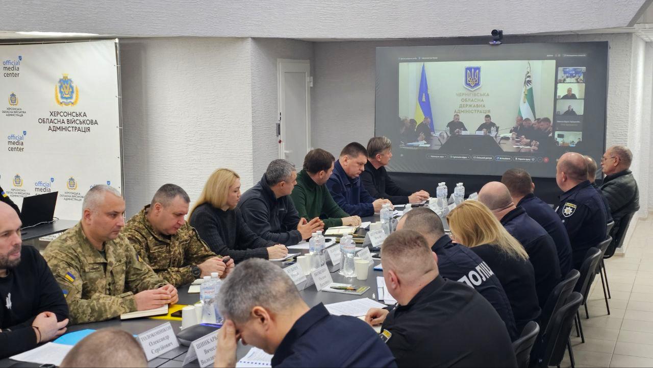 Група українських посадовців, правоохоронців та військових бере участь у нараді за великим столом. На задньому плані видно екран із відеоконференцією з Чернігівською обласною державною адміністрацією. Стіна прикрашена логотипом Херсонської ОВА.