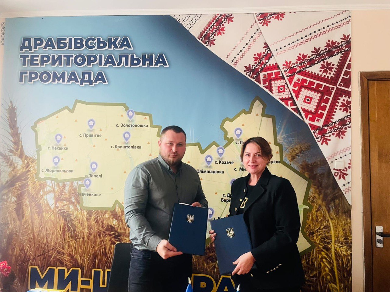 Двоє людей — чоловік і жінка — стоять поруч у приміщенні та тримають у руках темно-сині папки з гербом України. Чоловік, що стоїть ліворуч, має бороду та одягнений у сіру сорочку. Жінка, що стоїть праворуч, одягнена у чорний піджак. За ними на стіні висить велика карта, яка ілюструє Драбівську територіальну громаду та позначає назви її сіл. Частина стіни праворуч прикрашена елементами української вишиванки.