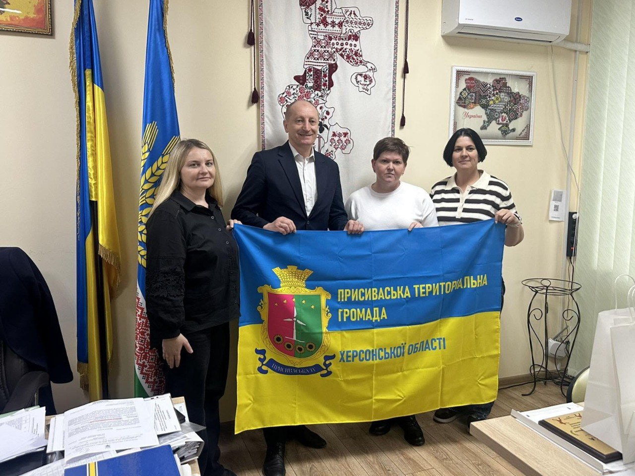 Четверо людей — двоє чоловіків і дві жінки — стоять разом у приміщенні та тримають розгорнутий прапор Присиваської територіальної громади Херсонської області. Чоловік, що стоїть у центрі, одягнений у чорний костюм і білу сорочку. Жінки одягнені у чорний, білий та смугастий одяг. Ліворуч від них видно прапори України.
