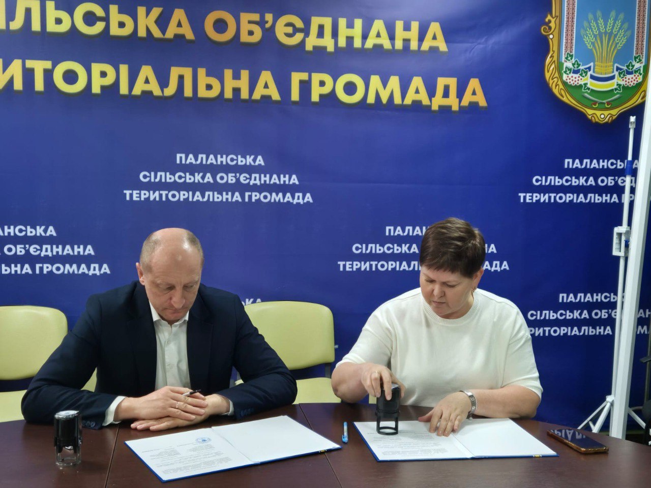 Двоє людей сидять за столом на тлі синього банера з біло-жовтим написом: ПАЛАНСЬКА ОБ'ЄДНАНА СІЛЬСЬКА ТЕРИТОРІАЛЬНА ГРОМАДА. Жінка праворуч, одягнена у білу футболку, ставить печатку на документі, який лежить перед нею. Чоловік ліворуч, одягнений у темно-синій костюм і білу сорочку, уважно спостерігає, склавши руки на столі. На задньому плані видно багаторазове повторення назви громади.