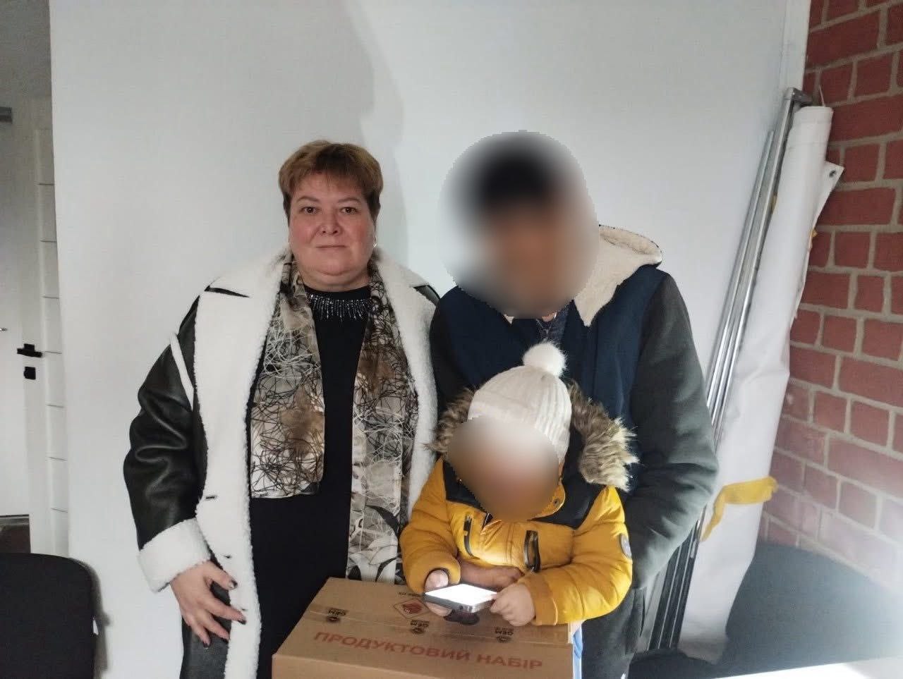 Жінка у світлому пальті стоїть поряд з двома дітьми, обличчя яких приховані мозаїкою. Жінка та старша дитина стоять біля картонної коробки з написом: ПРОДУКТОВИЙ НАБІР. Молодша дитина в жовтій куртці та білій шапці тримає в руках мобільний телефон.