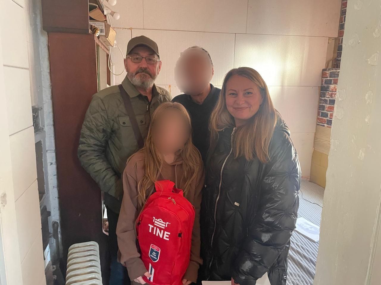 Чотири людини стоять разом, імовірно, у дверному отворі. Ліворуч — чоловік у кепці та зеленій куртці. Поруч із ним — дівчинка з довгим волоссям та червоним рюкзаком TINE. Праворуч — жінка в блискучій чорній куртці, яка посміхається. Між ними стоїть ще одна людина із закритим обличчям.