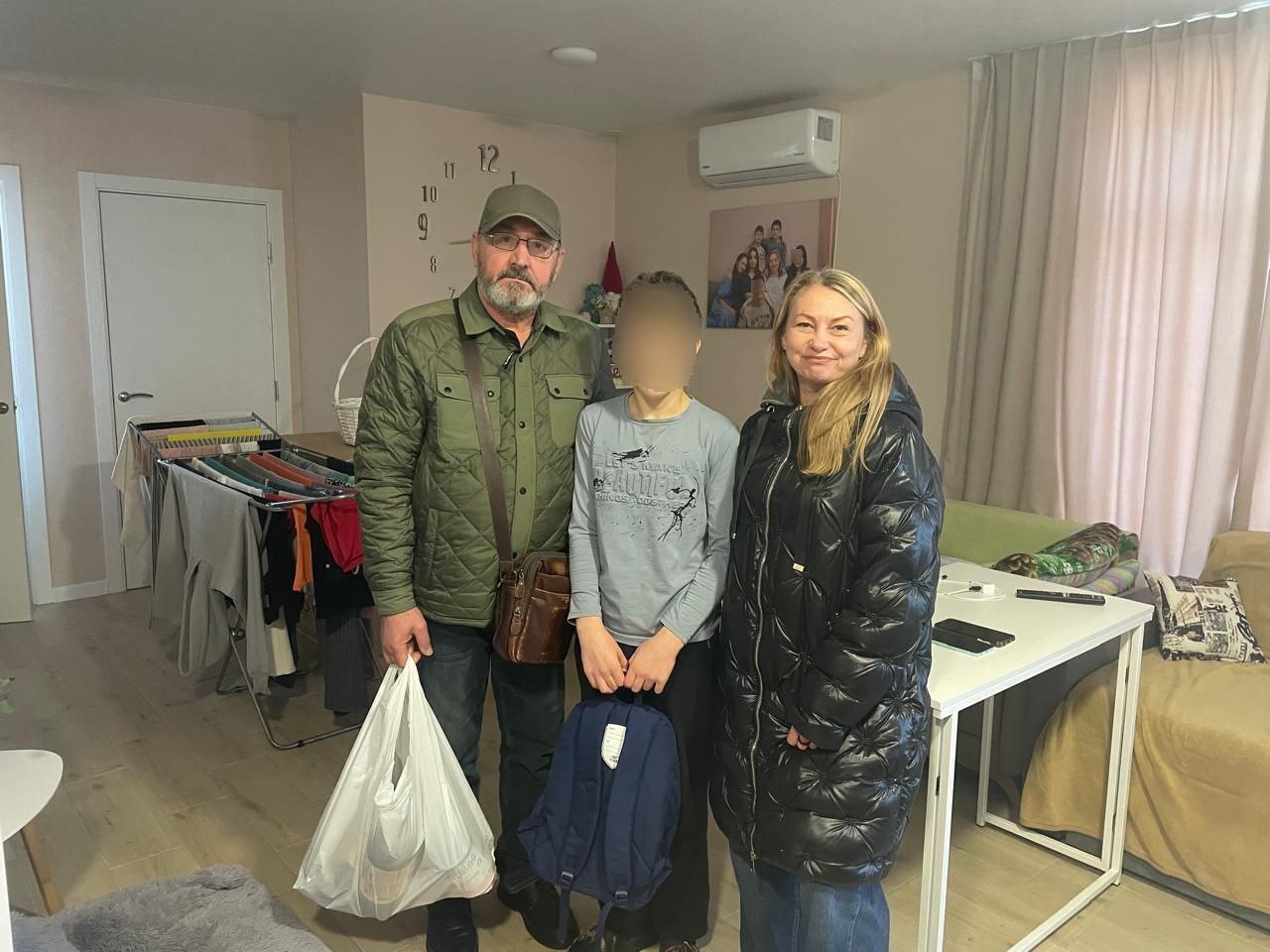 Чоловік у зеленій куртці, хлопчик у світшоті та жінка у чорній куртці стоять у світлій кімнаті. Чоловік тримає білий пакет, хлопчик — синій рюкзак. На обличчя дитини накладено мозаїку для збереження приватності.