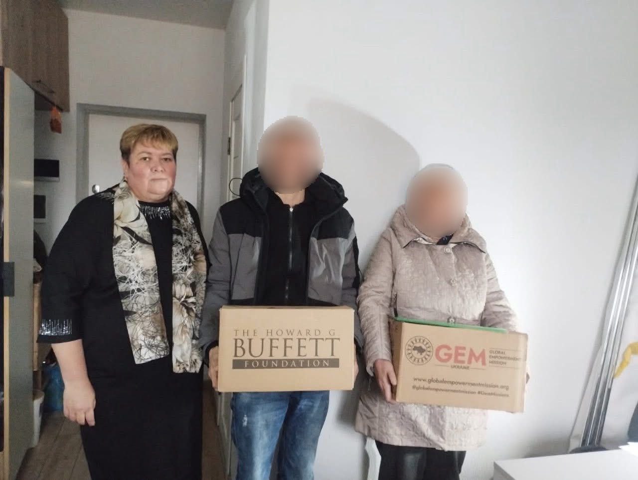 Троє людей тримають картонні коробки з гуманітарною допомогою в приміщенні. Ліворуч стоїть жінка в чорній сукні та хустці. Двоє інших людей (з розмитими обличчями) тримають коробки: одна від The Howard G. Buffett Foundation (Фонд Говарда Г. Баффета), інша від GEM - Global Empowerment Mission.