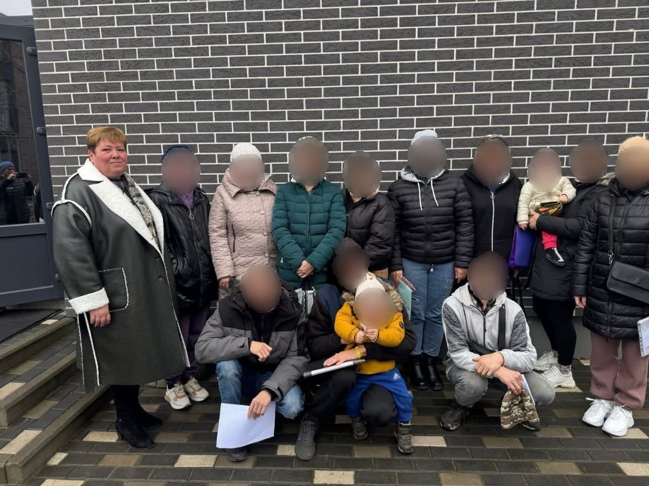 Група з 14 людей, дорослих і дітей, стоїть і сидить перед стіною з чорної цегли. Жінка у чорно-білому пальті стоїть ліворуч, а решта людей, включаючи дітей, розташовані ззаду і внизу. Обличчя більшості осіб приховані мозаїкою для збереження приватності.