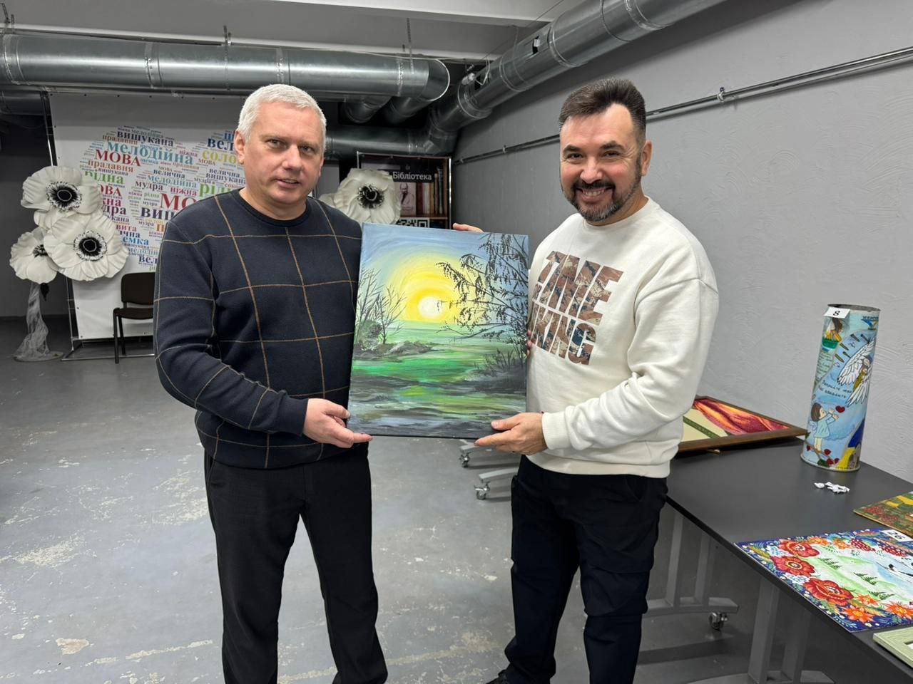 Двоє чоловіків, що стоять у приміщенні галереї чи лофту, тримають картину з пейзажем. Чоловік ліворуч має коротке сиве волосся і одягнений у темний светр у клітинку. Чоловік праворуч має темну бороду і одягнений у білий светр з написом: TIME KING. Вони дивляться прямо на камеру, тримаючи картину із зображенням заходу чи сходу сонця над болотистою місцевістю з темними деревами. На задньому плані видно сіру стіну та столи з іншими виставленими предметами мистецтва.