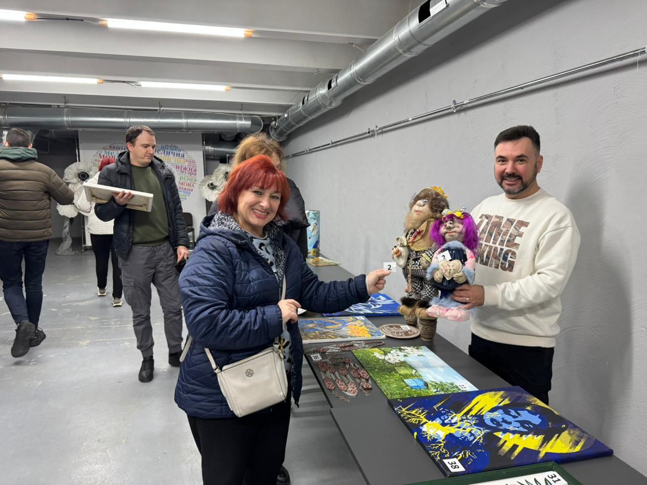 На виставці або ярмарку жінка з рудим волоссям у синій стьобаній куртці та світлій сумці через плече вказує пальцем на дві великі текстильні ляльки-обереги, які тримає усміхнений чоловік у кремовому светрі. Перед ними на довгому чорному столі розкладені витвори мистецтва, включаючи картини з абстрактними жовто-синіми елементами та пейзажні роботи. На задньому плані, у приміщенні з сірими стінами та відкритими трубами під стелею, видно інших відвідувачів, які розглядають експонати, зокрема чоловіка в сірих штанах та оливковій куртці, який тримає коробку.