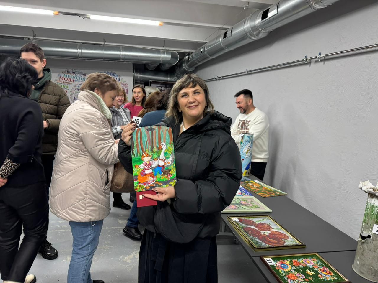 На виставці жінка з коротким світло-каштановим волоссям у чорній стьобаній куртці та спідниці тримає руками невелику картину з яскравим зображенням двох гусей та лисиці в лісі. Вона посміхається, дивлячись прямо в кадр. На задньому плані видно інших відвідувачів у повсякденному одязі та чорний стіл з виставленими художніми роботами, зокрема картинами, обрамленими бісером.