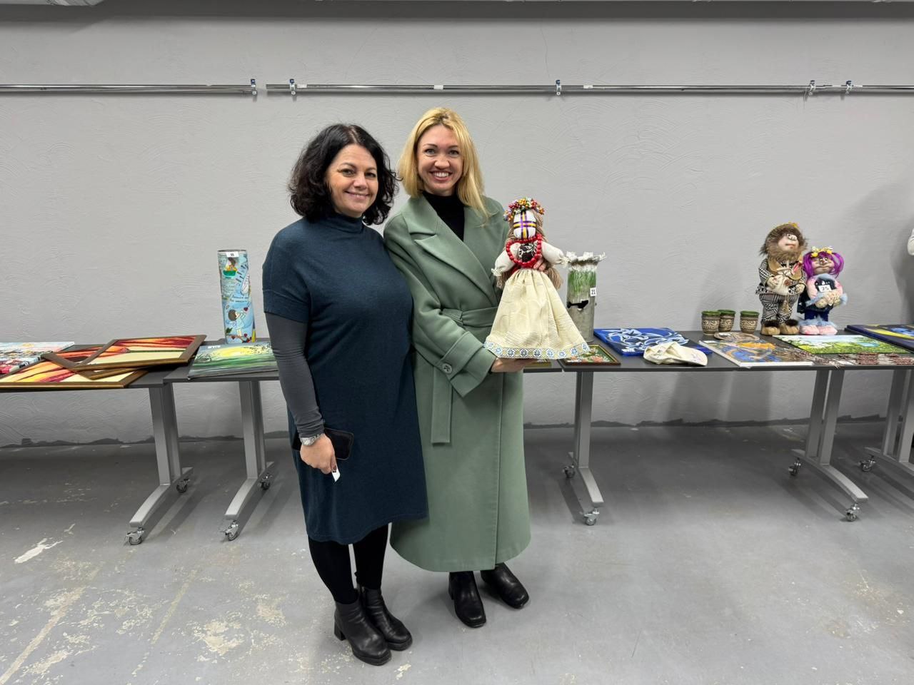Дві жінки стоять поруч і дивляться в камеру на тлі сірої стіни. Жінка ліворуч — із темним кучерявим волоссям, одягнена у темно-синю сукню. Жінка праворуч — зі світлим волоссям, одягнена у довге пальто кольору м'яти. Вона тримає в руках велику традиційну українську ляльку-мотанку. На столі позаду них виставлено різні предмети мистецтва та вироби, включаючи картини, скляні банки та інші ляльки чи фігурки.