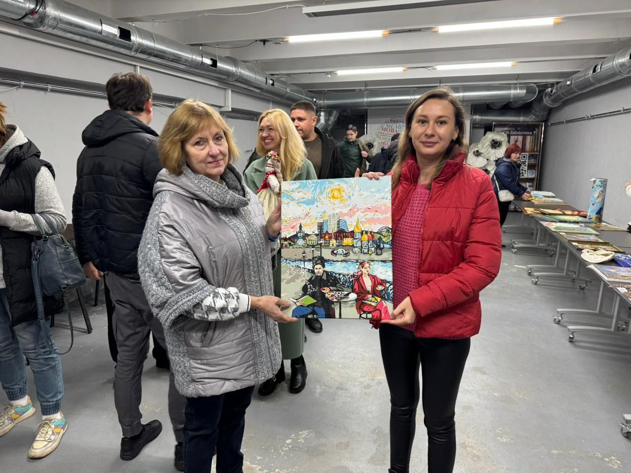 Дві жінки, що стоять у приміщенні серед інших людей, тримають велику картину. Жінка ліворуч — у блискучій сірій куртці та сірому шарфі, а жінка праворуч — у червоній куртці та чорних штанах. Вони обоє дивляться прямо на камеру, тримаючи картину горизонтально. На картині зображено міський пейзаж, а на передньому плані зображено Тараса Шевченка та жінку, які сидять і займаються мистецькою діяльністю.