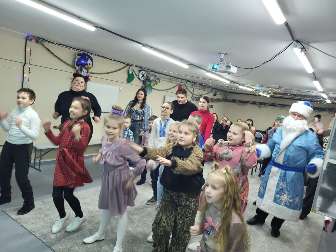 Група дітей та дорослих, включаючи персонажа Святого Миколая в блакитній шубі, весело танцюють і рухаються в приміщенні, прикрашеному гірляндами та повітряними кульками. Це виглядає як дитячий захід.