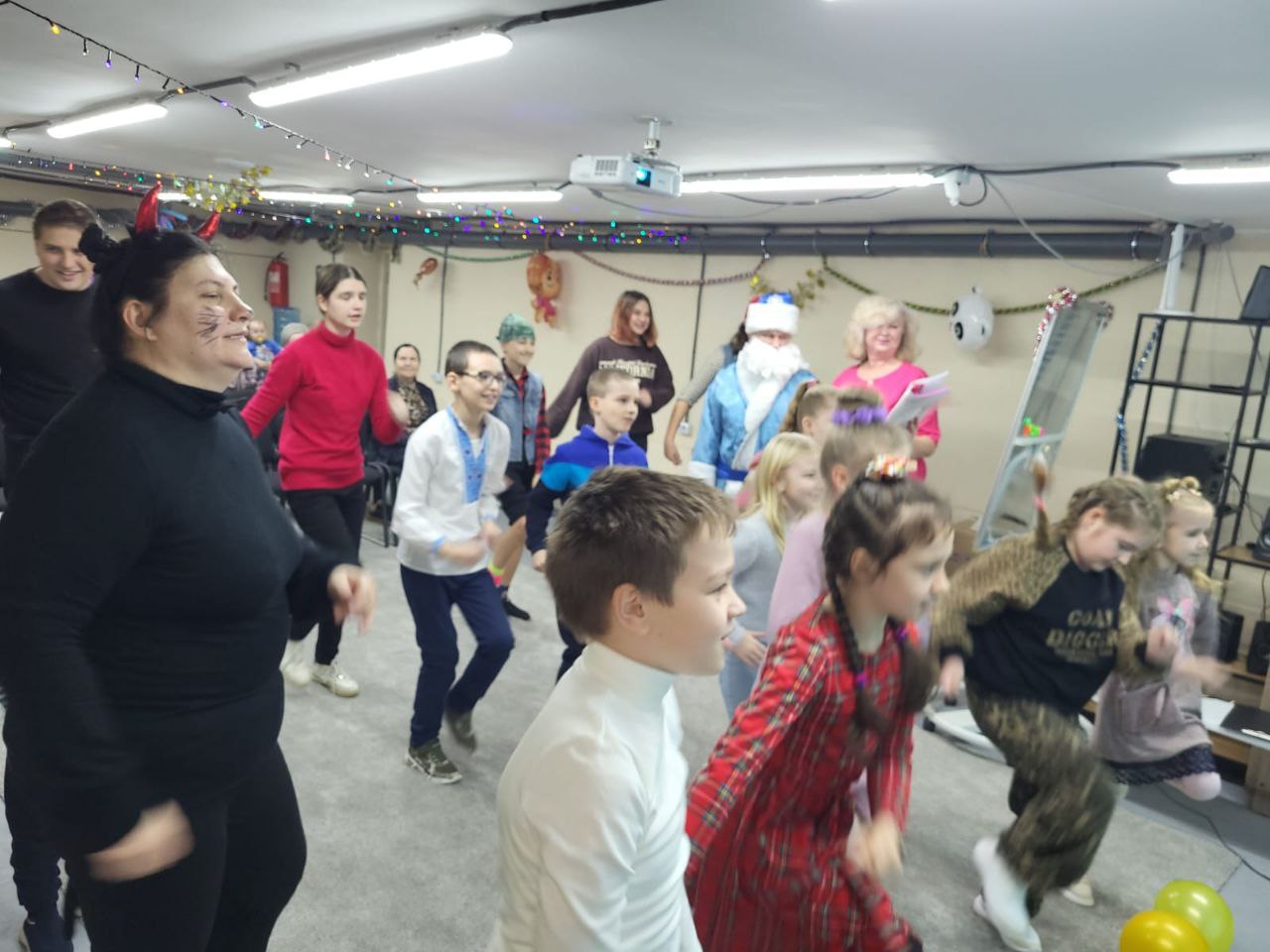 На фото зображена група людей, переважно дітей та підлітків, які активно танцюють або роблять рухи під час святкового заходу у приміщенні. Серед них є дорослі, включаючи жінку в чорному з намальованими вусами та ріжками, та персонажа Святого Миколая у блакитному вбранні. Приміщення прикрашене гірляндами.