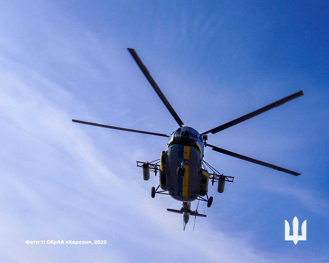 На фото показано військовий вертоліт у кольорах прапора України (синій і жовтий), що летить низько в небі, знятий знизу. На задньому плані видно чисте блакитне небо. У нижньому лівому куті розташований підпис: Фото: 11 ОбрАА Херсон, 2025.