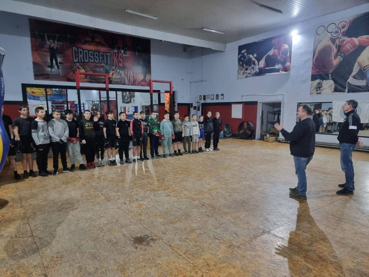 Група юних спортсменів (близько 20 хлопців та дівчаток) стоїть у ряд у боксерському залі. Перед ними праворуч стоять двоє дорослих чоловіків, один з яких розмовляє. На стінах залу видно великі банери з написами: CROSSFIT KS та зображення боксерів.