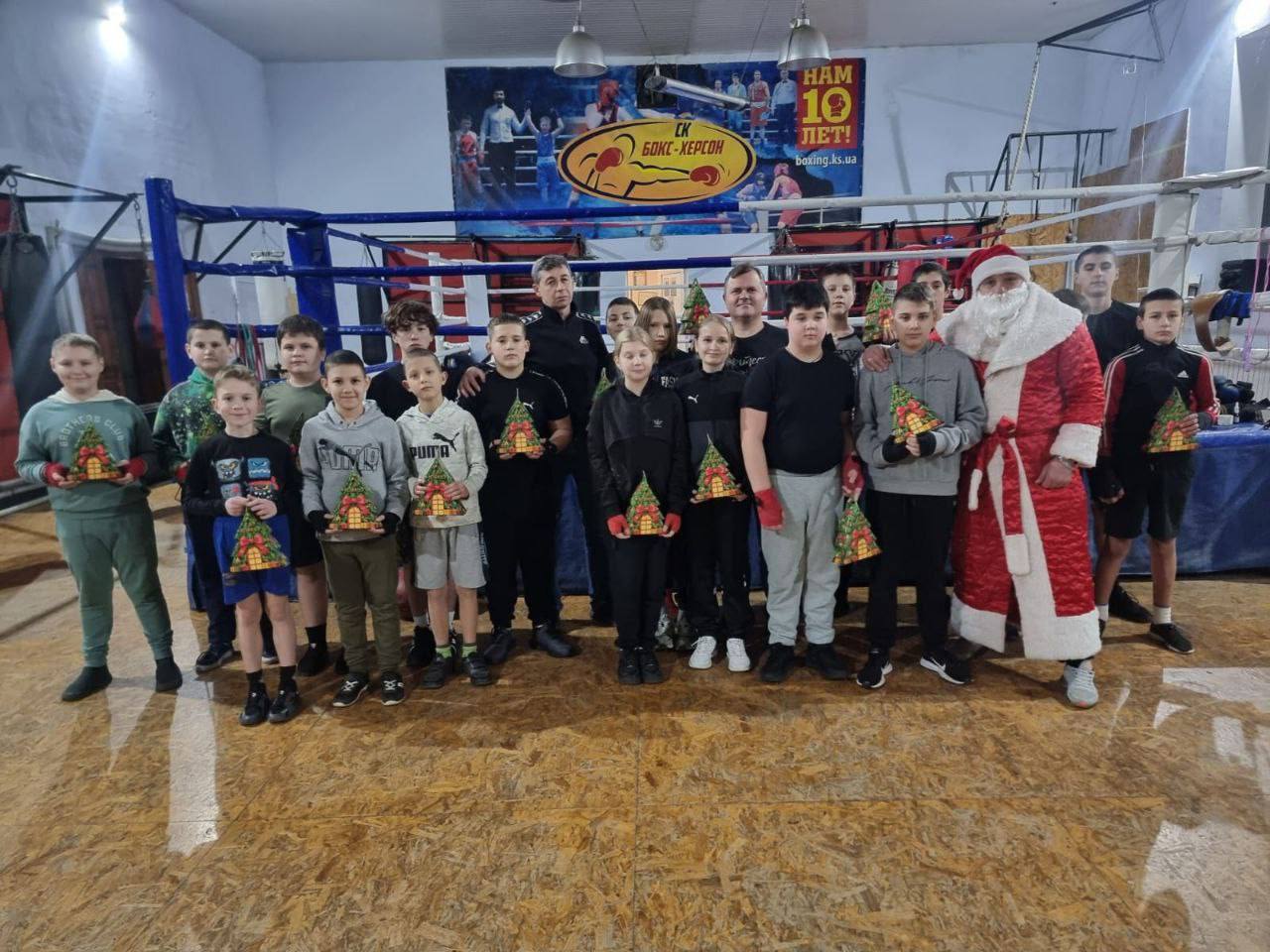Групова фотографія в боксерському залі. На знімку — близько 20  юних спортсменів: хлопців та дівчаток, кілька дорослих чоловіків (частина з яких тренери), які стоять у рингу або біля нього. Праворуч стоїть чоловік, одягнений як Святий Миколай і всі діти тримають у руках невеликі подарунки (упаковки у формі ялинки). На задньому плані видно банер СК Бокс-Херсон та напис: НАМ 10 ЛЕТ!