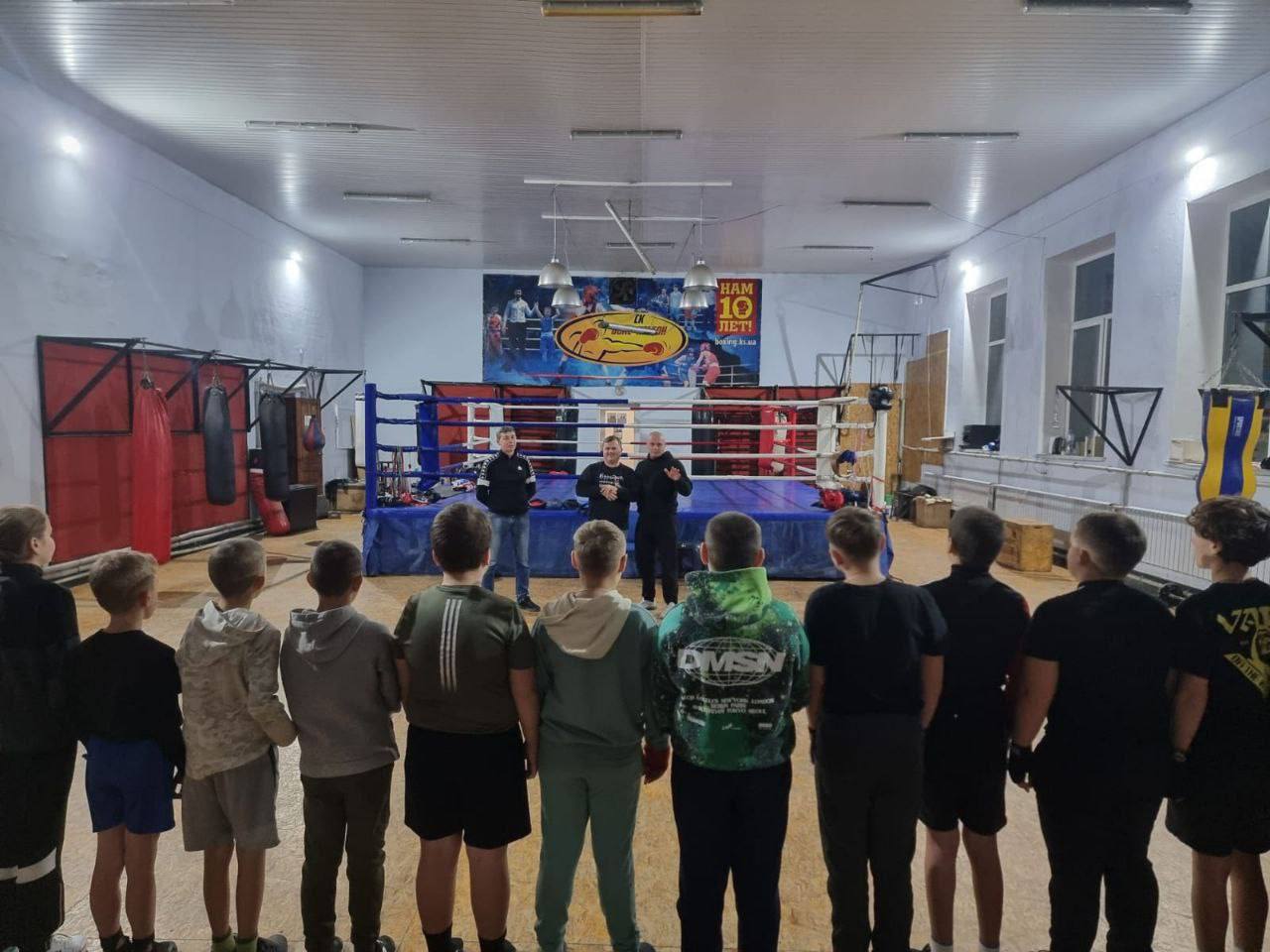 Група юних спортсменів (хлопців та дівчаток) стоїть спиною до камери, вишикувавшись у ряд у великому спортивному залі, дивлячись на боксерський ринг. У рингу стоять троє дорослих чоловіків, один з яких розмовляє. На задньому плані над рингом розташований банер із зображеннями боксерів та написом: НАМ 10 ЛЕТ!
